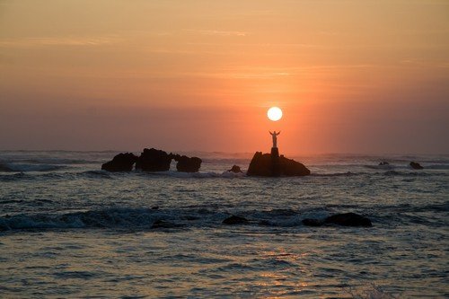 The sun sets over a statue of Jesus out in the ocean in El Salvador